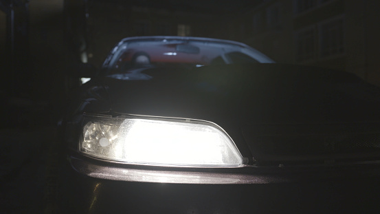 Close up for a headlight of a parked car being turned off at night. Details of a vehicle, front bumper and car headlight standing near the house.