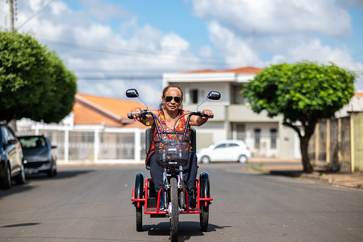 Portraits of a Black senior lady with much vitality and joy, wearing cool sunglasses and hanging out in a small city with her electric tricycle. She is riding on  calm streets with some modest and soporific houses around her, in a summer day with blue sky with clouds.