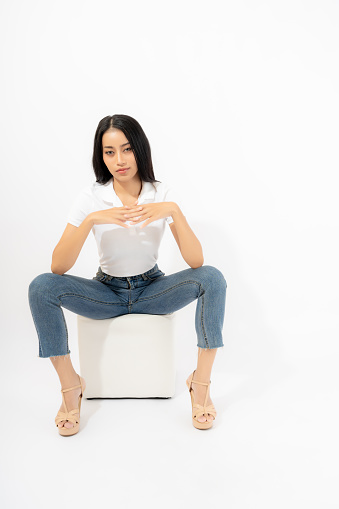 Beautiful confident young asian woman in jeans high heel collar shirt sitting on sofa cube isolated on white background Studio shot Attractive girl looking at camera Full length of Asia female posing