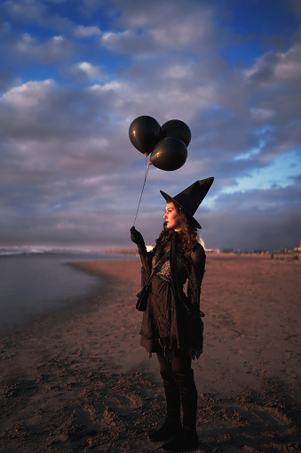 woman dressed as a witch on the shore of the beach holding black balloons