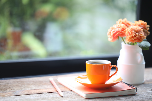 Orange roses and small coffee cup and notebook pencil on wooden in front of windows