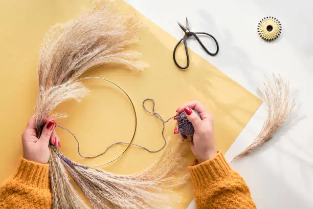 Hands making dried floral wreath from dry pampas grass and Autumn leaves. Hands in sweater tie decorations to metal frame. Flat lay on white yellow table, sunlight with long shadows.