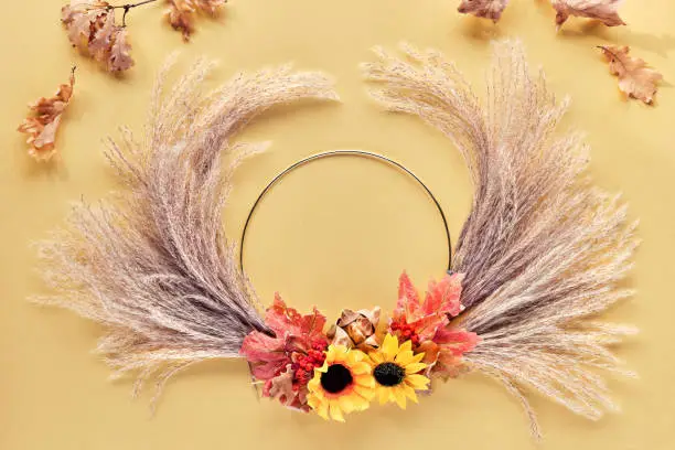 Hands making dried floral wreath from dry pampas grass and Autumn leaves. Hands in sweater tie decorations to metal frame. Flat lay on white yellow table, sunlight with long shadows.