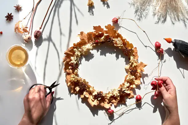 Hands making dried floral wreath from dry Autumn leaves and Fall berries. Hands with manicured nails fixing decorations with glue gun. Flat lay on white table.