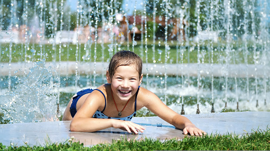smiling, happy eight year old girl in swimsuit having fun in splashes in street city fountain, outdoors, in park, summer, hot sunny day during vacation. High quality photo