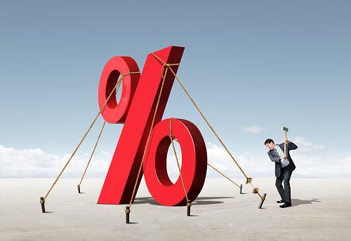 A man swings an axe at the ropes that restrain a tall red percent sign. The ropes are attached to stakes in the ground conveying concept of artificially low rates restrained by Federal Reserve policies.  The rope he is taking aim at is frayed and on the brink of being completely severed.