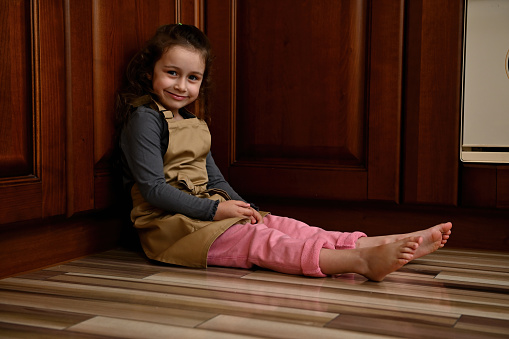 Beautiful preschool age child, a charming European little girl, cute baker confectioner wearing a beige chef's apron, sitting barefoot on the kitchen floor near a cupboard, smiling looking at camera