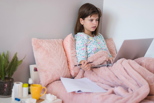 Serious school girl sitting in bed when sick and finishing homework with laptop and a notepad