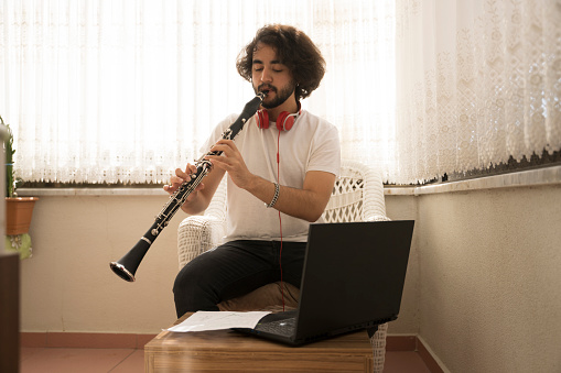 Young man playing the clarinet on the balcony of the house.\nyoung long-haired boy playing a woodwind instrument at home.