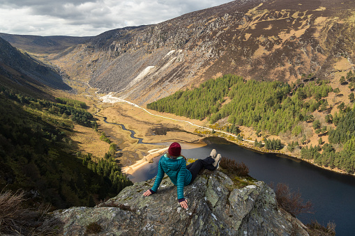 Woman Hiker Sitting on a Rock on the Top of a Mountain admiring the lake and landscape in Glendalough Wicklow Mountains