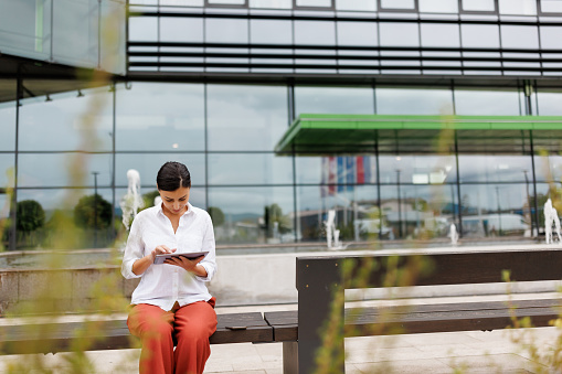 Beautiful young businesswoman sitting on bench in front of office building and using digital tablet