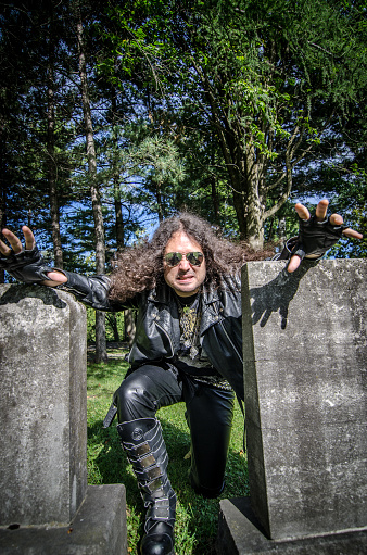 Heavy metal musician all dressed up in black leather with sunglasses and long curly hair is crawling between two tombstones in a cemetery during day of summer