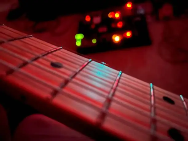 This is a closeup blurred shot of a guitar fretboard with a guitar pedal board in the background and is also blurred