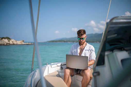 Man on vacation with his sailboat. He is taking some time to work remotely on his laptop.