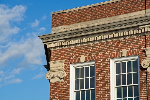Ornate concrete cornice at roof line above decorative ionic column detail on corner of red brick commercial building in Lexington Massachusetts. Bright blue skies with cumulus clouds.