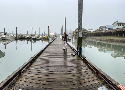 On August 25 ,2022 on Resurrection Bay pier in Seward Alaska a woman is walking along the waterfront viewing the many boats harbored at the slots in the pier.  The day was cold raining and foggy.