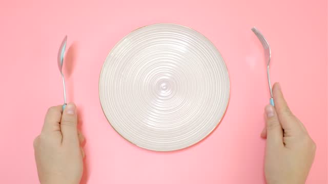 Female hands holding a fork and spoon next to an empty plate on a pink background close-up top view.