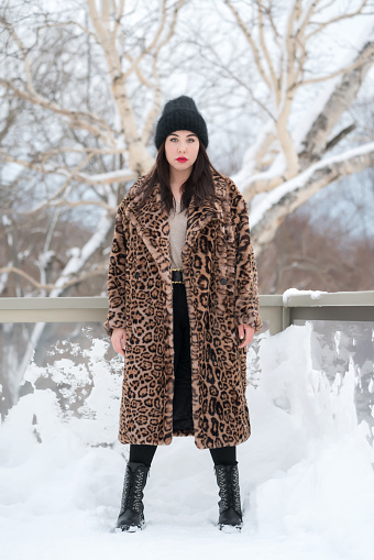 Full length portrait of plus size young woman in fur coat with leopard pattern, knitted hat, black jeans and high boots. Brunette woman standing straight, legs at shoulder level, looking at camera. Part of a series of photos.