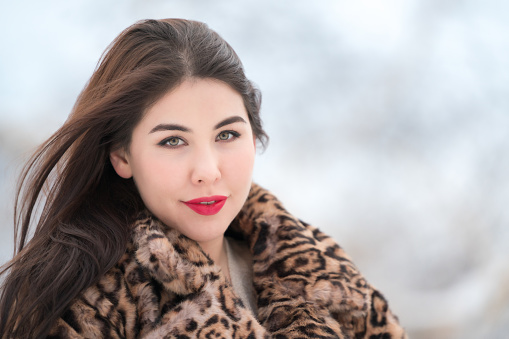 Portrait of happy smiling brunette woman with long hair and red plump lips, dressed in faux fur coat with leopard pattern and looking at camera. Winter portrait of elegant young woman outdoors. Part of a series of photos.