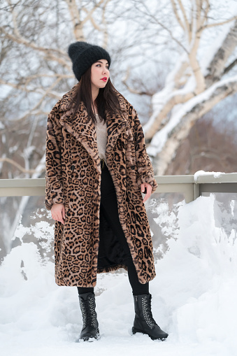 Full length view of young adult female in fur coat with leopard pattern, knitted hat, black jeans and high boots. Winter portrait of charming plus size brunette woman standing in snow and looking away. Part of a series of photos.