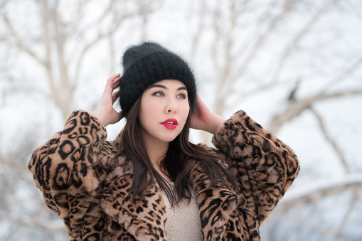 Winter portrait of young adult female with long hair and red plump lips, dressed in fur coat with leopard pattern and black knitted hat. Brunette woman raised hands and straightens hat, looking away. Part of a series of photos.
