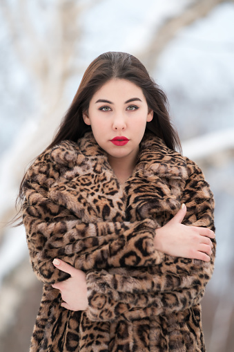 Portrait of brunette young woman with long hair and red plump lips, dressed in faux fur coat with leopard pattern looking at camera and hugging herself. Winter portrait woman outdoors in cold weather. Part of a series of photos.