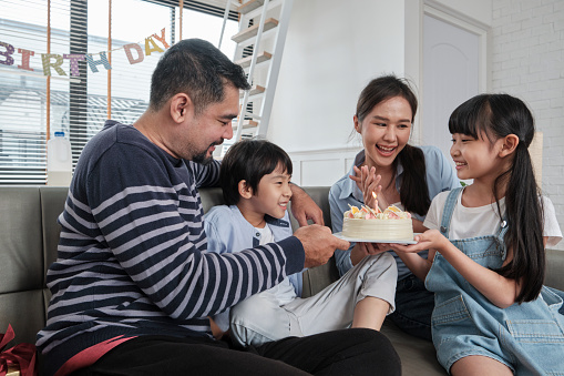 Happy Asian Thai family, young son is surprised with birthday cake, gift, blows out candle, and celebrates party with parents together in living room, wellbeing domestic home event lifestyle.