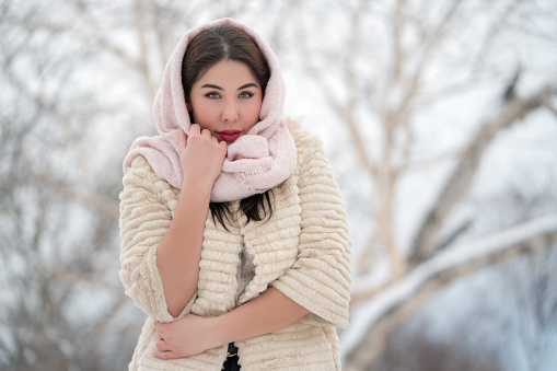 Authentic brunette young woman dressed in white three-quarter sleeve fur coat and light pink scarf on head looking at camera on winter day outdoors. Shallow depth of field, selective focus foreground. Part of series of photos