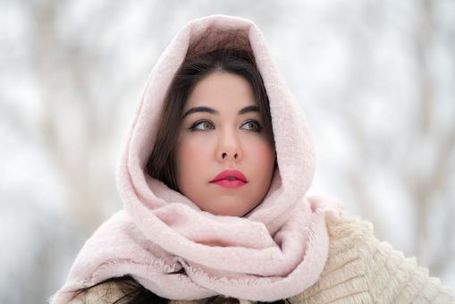 Portrait of thoughtful brunette young woman dressed in white fur coat and light pink scarf on head looking away. Romantic adult female model on winter day outdoors. Selective soft focus on foreground. Part of series of photos