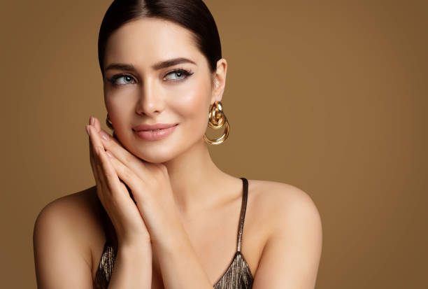 frau schönheit mit glatter haut make-up und goldenem schmuck. schönes mädchen mit perfekten lippen und augen-make-up, das händchen unter dem kinn hält. elegantes modellporträt mit goldenem ohrring lächelnd - fashion model stock-fotos und bilder