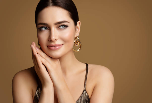 woman beauty with smooth skin make up and golden jewelry. beautiful girl with perfect lips and eye makeup holding hands under chin. elegant model portrait with gold earring smiling - fashion woman imagens e fotografias de stock