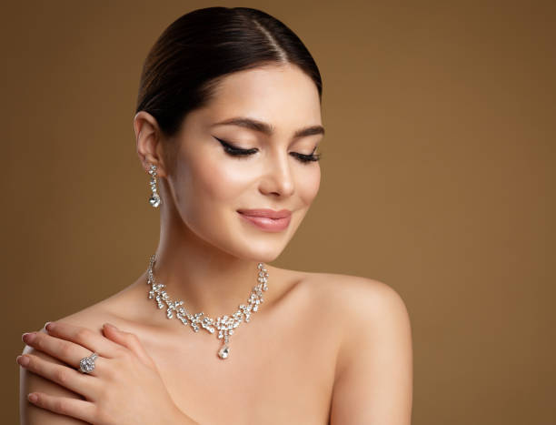 Beauty Model in Wedding Jewelry Set. Elegant Woman in Necklace with Earring and Ring. Beautiful Girl with perfect Eyeliner Make up and smooth Skin over Beige Background stock photo