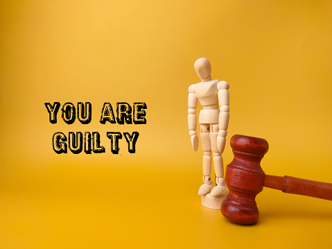Wooden mannequin and gavel with the word YOU ARE GUILTY on yellow background.