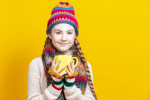 Winsome Teenage Girl in Warm Seasonal Hat and Knitted Scarf Holding Big Yellow Cup While Drinking tea or Coffee Against Yellow Background. Horizontal Composition