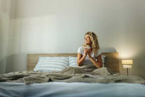 Young happy woman enjoying while day dreaming during morning coffee time on a bed in bedroom. Copy space.