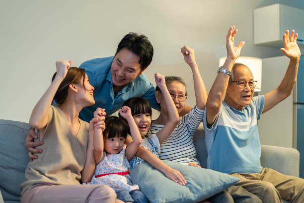 asian family cheering and watching football on television at home.  muti-generation, senior old couple and young parents sit on sofa with little children looking at tv and feeling happy in living room - gamer watching tv adult couple imagens e fotografias de stock