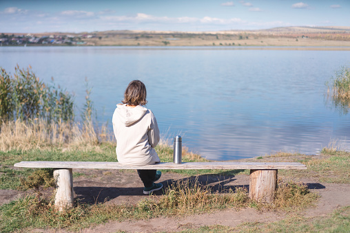 young woman drinking tea  from thermos near by a lake