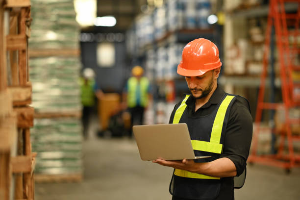 istock Warehouse manager wearing hardhats and reflective jackets using laptop for checking stock and order details in warehouse. 1427500453