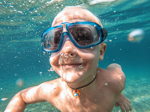 Cute blonde boy wearing goggles and holding his breath underwater. He is swimming and diving at the sea.