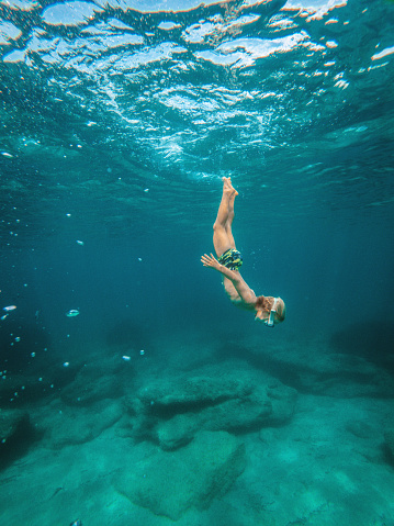 Young caucasian explorer snorkeling underwater at the sea. He is moving down in vertical position. He is wearing swimwear and goggles.