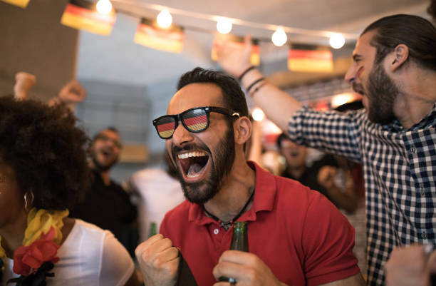 Group of young multi-ethnic soccer fans cheering at the bar. Holding German team flags, with color paint on faces.