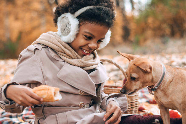 Cute African-American girl eats a croissant at a picnic with a dog in an autumn park.Diversity,autumn concept.