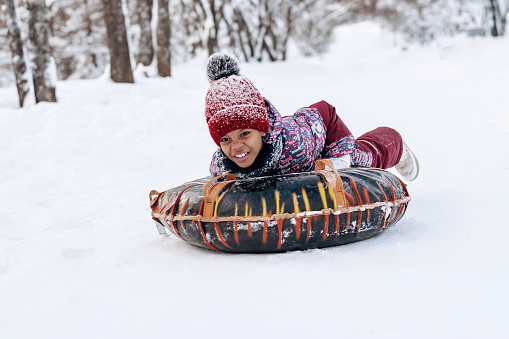 Happy little African-American girl in a red hat and jumpsuit rides on tubing in the winter park.Beautiful trees are covered with white snow.Winter fun,active lifestyle concept.Selective focus.