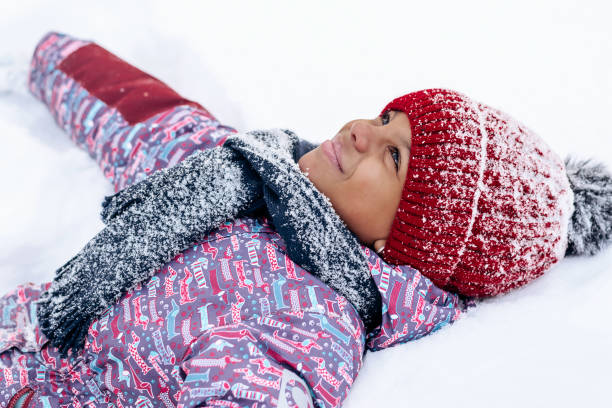 Happy little African-American girl in a red hat and jumpsuit is lying in the snow.Winter fun,active lifestyle concept