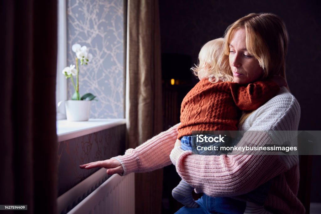 Mother With Son Trying To Keep Warm By Radiator At Home During Cost Of Living Energy Crisis Poverty Stock Photo