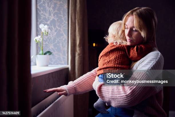 Mother With Son Trying To Keep Warm By Radiator At Home During Cost Of Living Energy Crisis Stock Photo - Download Image Now