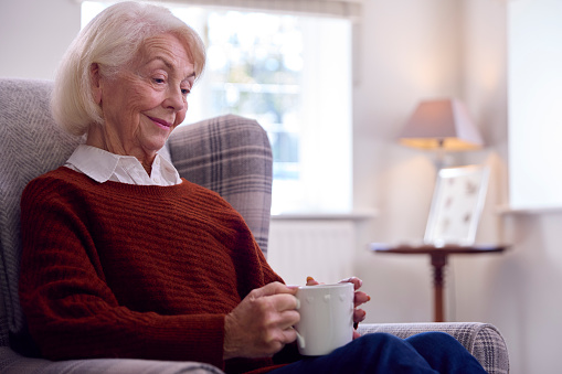 Senior Woman With Hot Drink Sitting In Armchair In Lounge At Home