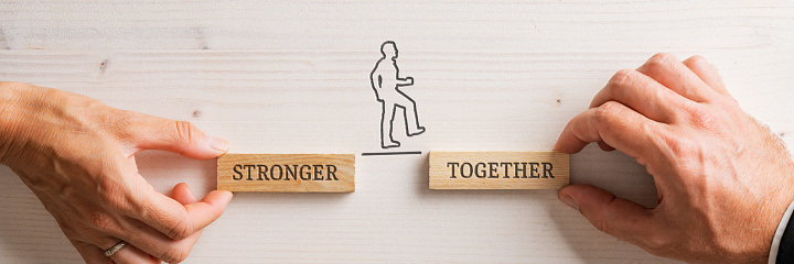 Hands of a businessman and businesswoman holding two wooden pegs with a Stronger together sign for a silhouetted businessman to walk confidently between the two. Wide view image.