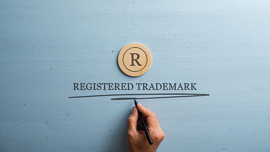 Letter R cut into wooden cut circle and male hand writing a Registered trademark sign under it. Over pastel blue wooden background.