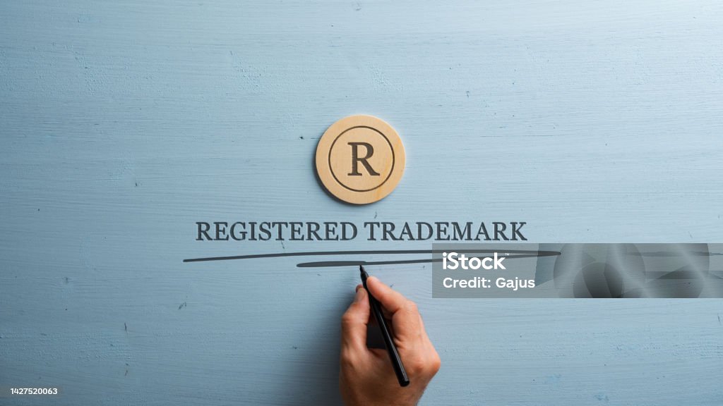 Letter R cut into wooden cut circle and male hand writing a Registered trademark sign under it Letter R cut into wooden cut circle and male hand writing a Registered trademark sign under it. Over pastel blue wooden background. Intellectual Property Stock Photo
